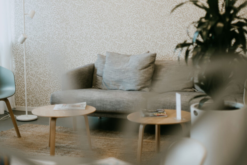 En modern och ljus lounge med en grå soffa, träbord och en grön växt i hörnet, sedd genom en suddig glasdetalj.