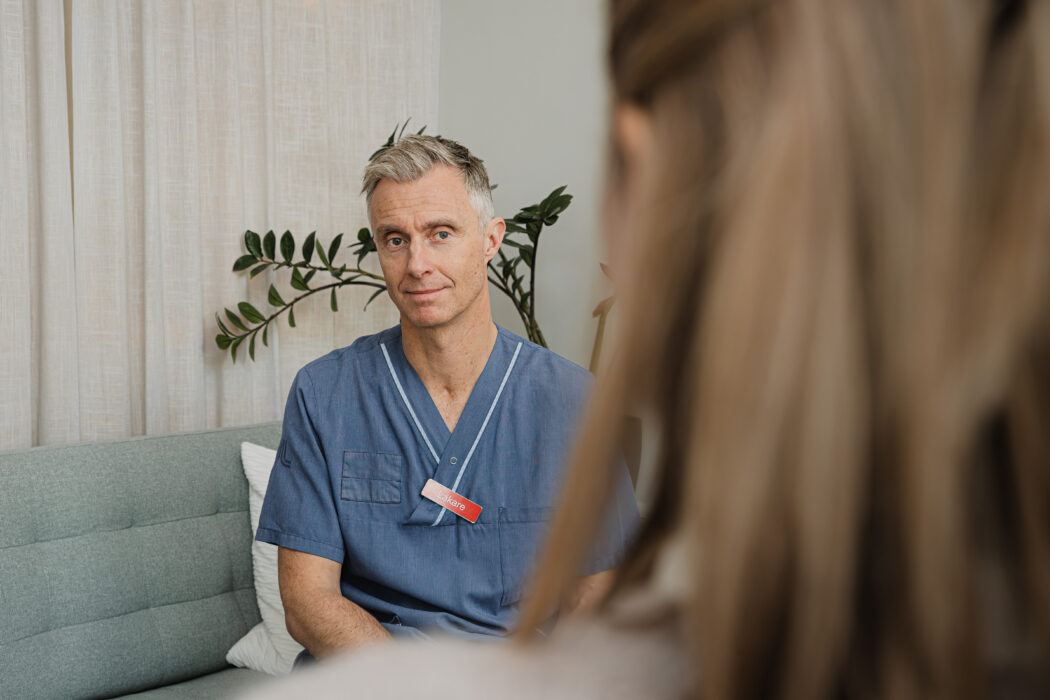 En person i blå vårdkläder sitter på en soffa, omgiven av växter och inomhusmiljö.