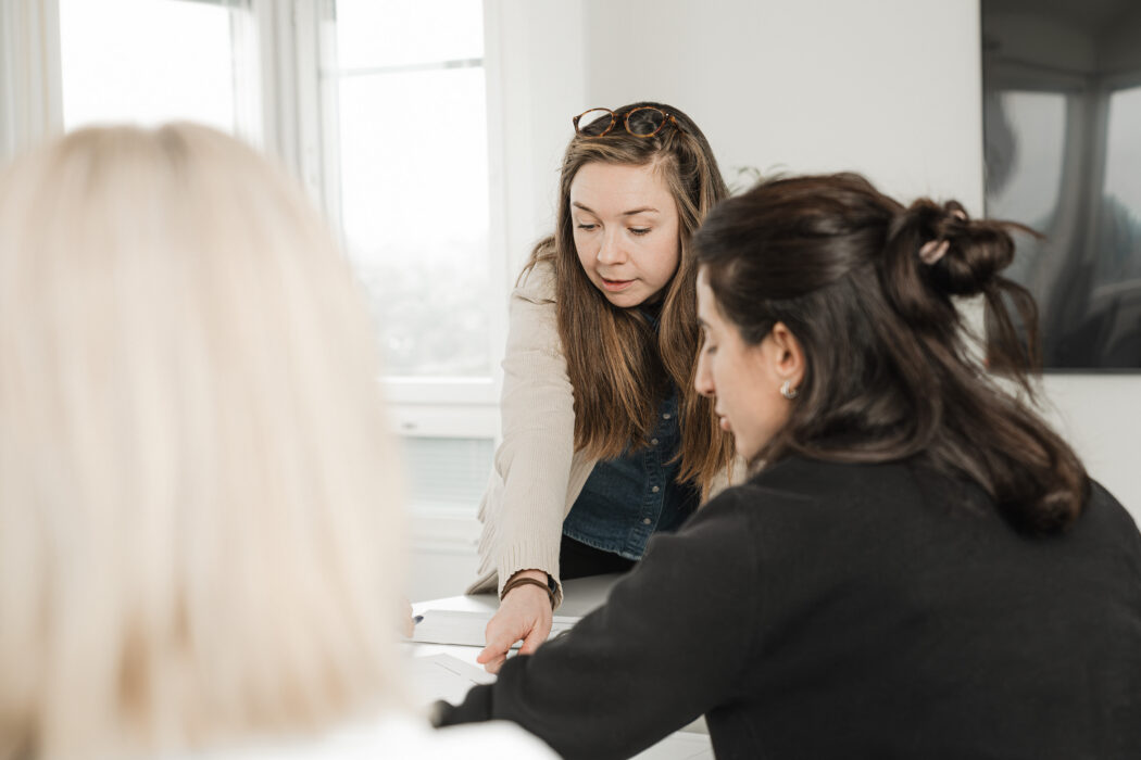 Tre personer sitter vid ett bord och verkar diskutera något. En person lutar sig framåt, medan de andra två sitter och lyssnar.