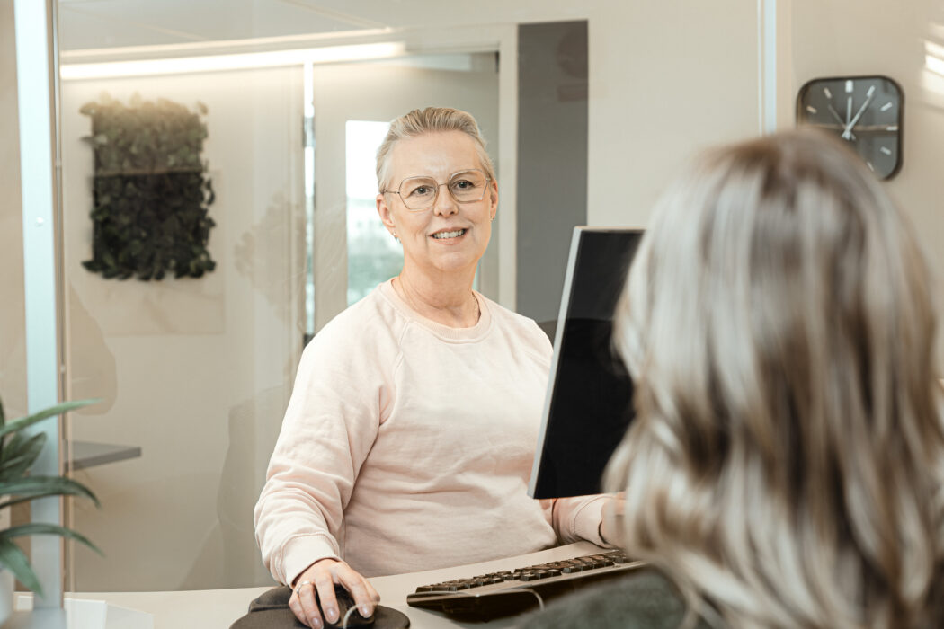 En person i ljus tröja sitter vid en dator och använder en mus, med en annan person med långt hår i förgrunden.