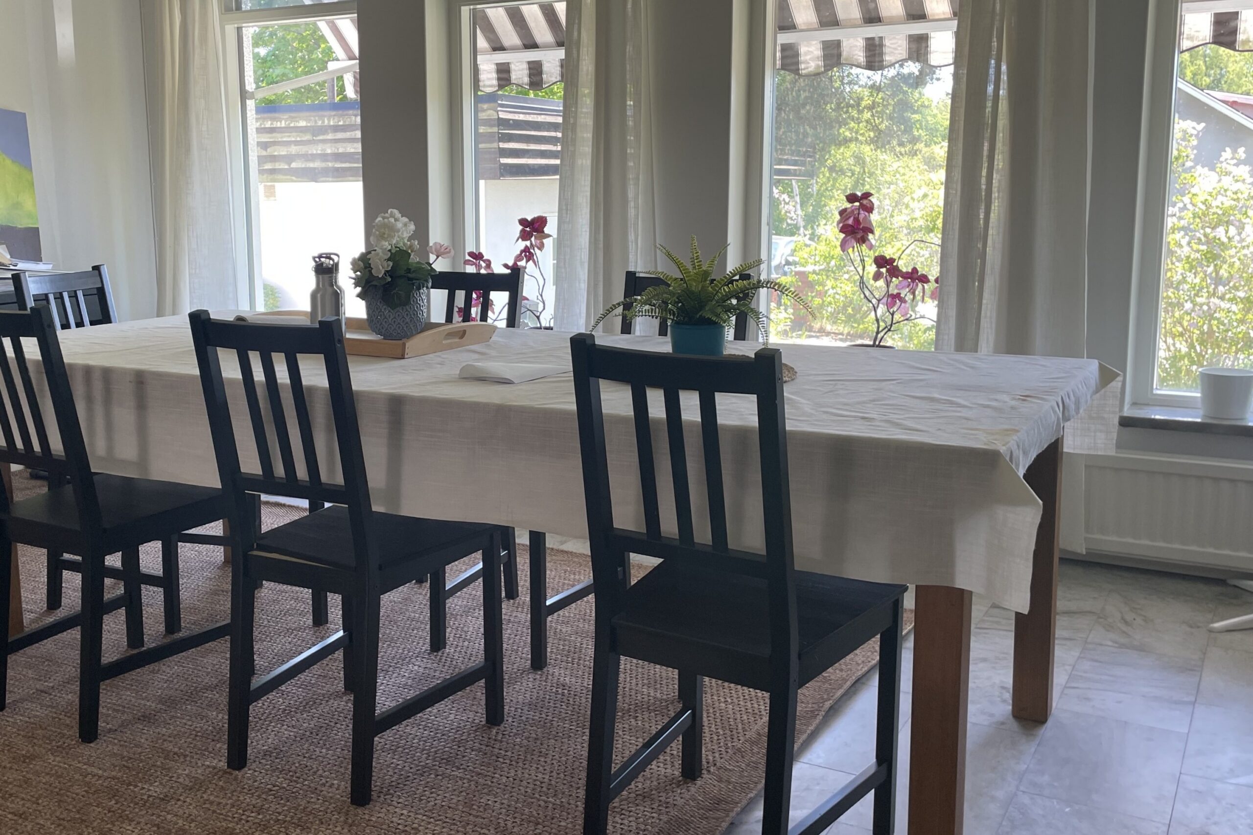 Ett matbord täckt med en vit duk, omgivet av svarta trästolar, dekorerat med en bricka med en vas med blommor och en vattenflaska samt en krukväxt; i bakgrunden syns stora fönster med utsikt över en trädgård med blommor och gröna växter.
